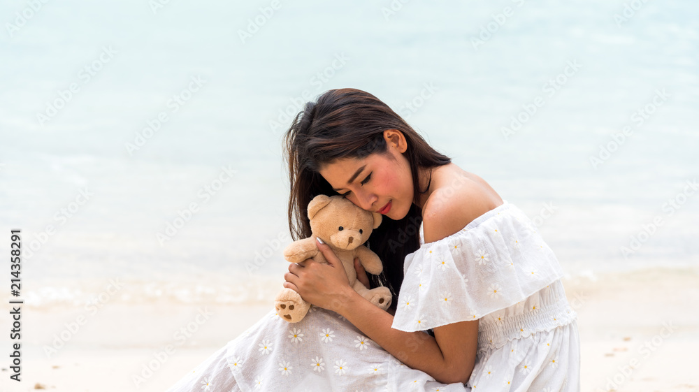 Asian girl in white dress sitting on the beach hugging a bear with love nostalgia.