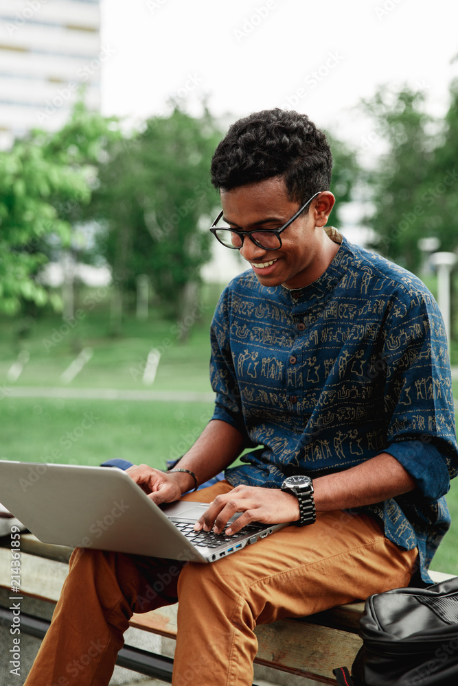 Young man university student online learning via laptop computer, sitting on a bench on a campus. Stylish hipster guy experienced freelancer working distance on notebook while resting in city park Young man university student online learning via laptop computer, sitting on a bench on a campus. Stylish hipster guy experienced freelancer working distance on notebook while resting in city park
