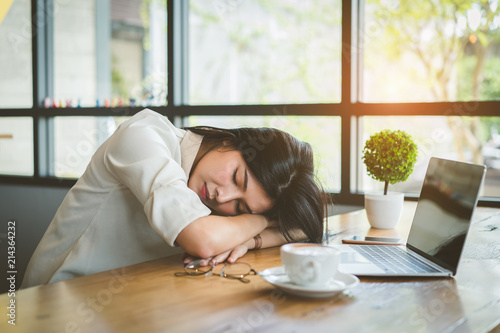 Freelancer asian businesswoman tired after working coffee shop her sleeping on workplace table near windows at evening with digital laptop computer and coffee break.