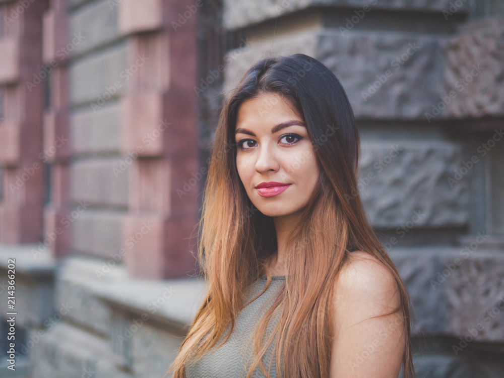 Portrait of young attractive girl looks into the camera with smile. Woman with oriental face, brown eyes and stylish ombre dyed long haistyle on architecture background. Street style.