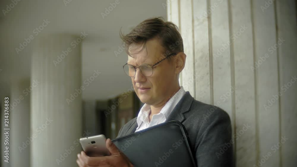 Businessman in the city. He wears a suit and briefcase. He looks through documents and talks on the smartphone Businessman in the city. He wears a suit and briefcase. He looks through documents and talks on the smartphone
