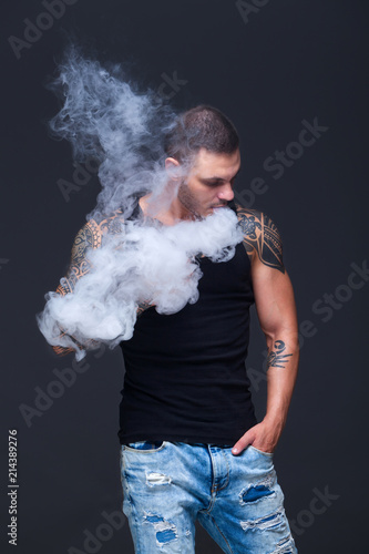 Vaper. The man with a muscular torso with tattoos smoke an electronic cigarette on the dark background