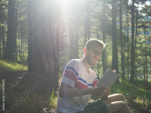 young handscome man in casual sitting on ground in the forest and writing poetry in notebook on a sunny day