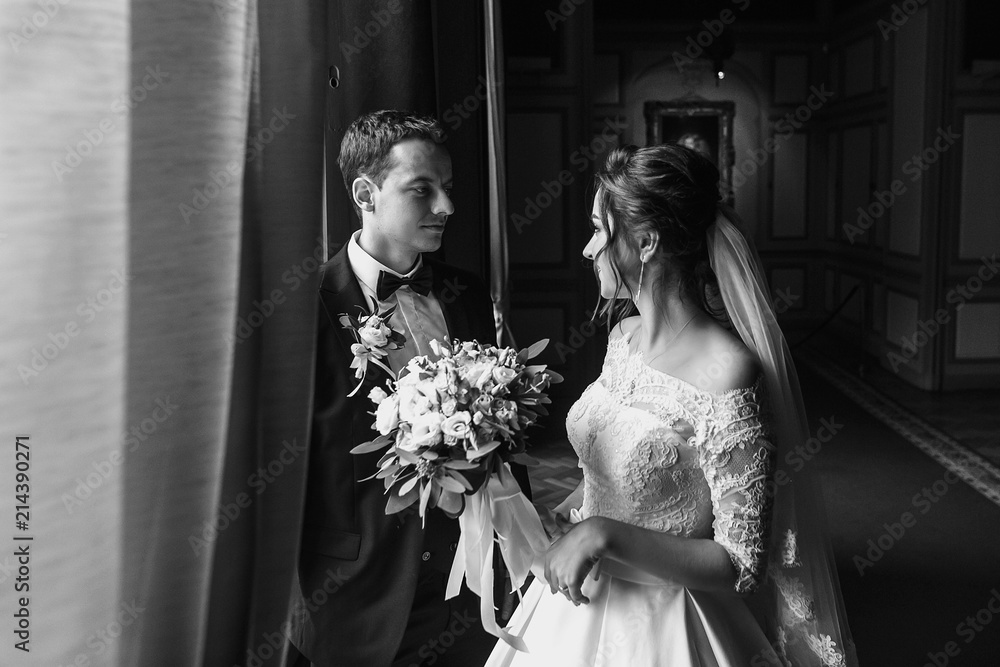 Φωτογραφία stylish groom looking at his gorgeous bride in stylish dress with amazing bouquet  in luxury room in hotel