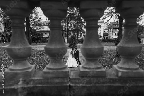 unusual view through columns at gorgeous bride and stylish groom walking at old castle in european park in autumn time. happy wedding couple embracing. romantic moment