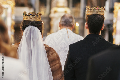 gorgeous bride and stylish groom in golden crowns, standing with priest in church during wedding ceremony. spiritual newlywed couple in holy matrimony. emotional moment