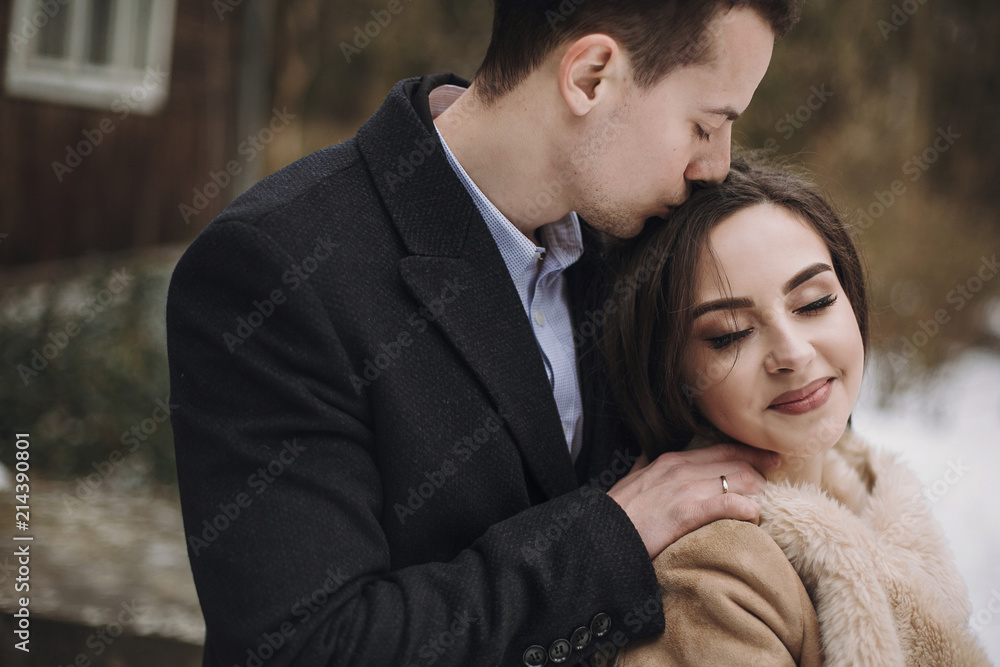 gorgeous bride in coat and stylish groom hugging at wooden house. wedding couple gently hugging and embracing in snowy park. barn wedding. romantic sensual moment of newlyweds gorgeous bride in coat and stylish groom hugging at wooden house. wedding couple gently hugging and embracing in snowy park. barn wedding. romantic sensual moment of newlyweds