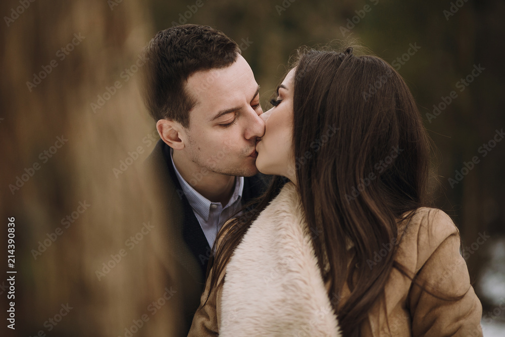gorgeous wedding couple kissing in winter snowy park. stylish bride in coat and  groom embracing under  trees in winter forest. romantic sensual moment of newlyweds