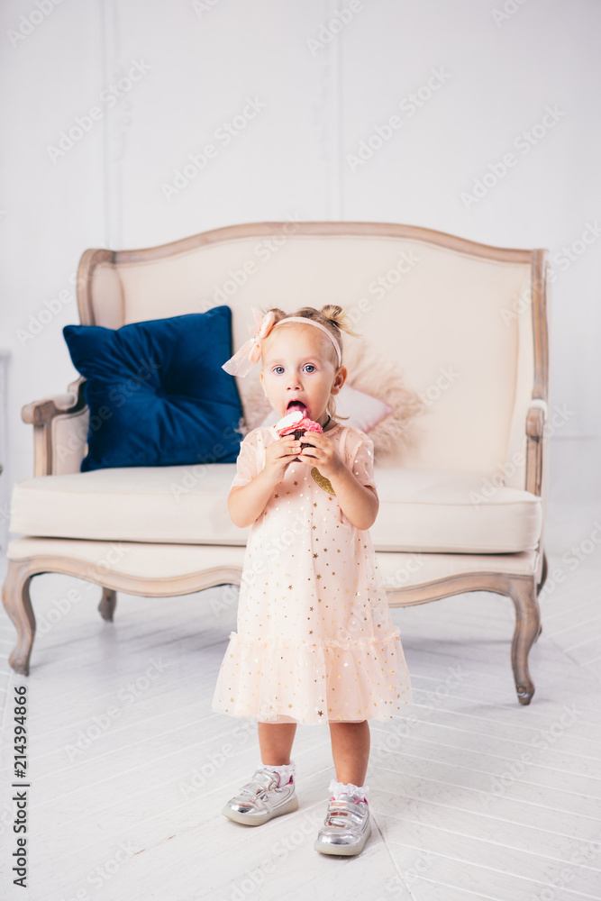 Obraz premium children's birthday. funny two-year-old Caucasian girl in pink dress standing to bedroom of house the background of couch and eating, holding cake dessert. Face and mouth smeared, smeared with cream