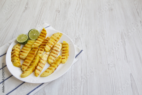 Grilled pineapple slices with lime on white plate over white wooden background, side view. Summer food. Idea for snack. Copy space and text area.