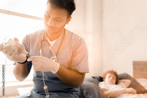 My profession. Competent pediatrician bowing head while looking at bottle with medicine and sitting on the foreground