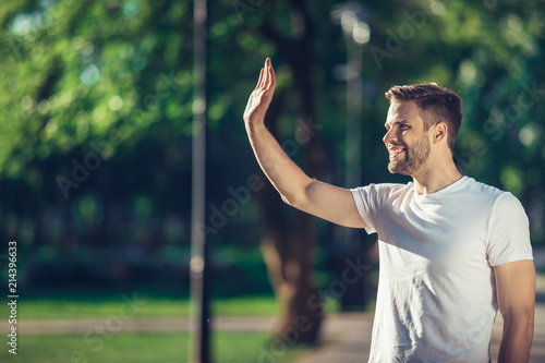 Side view of delighted male standing outdoors and waving hand to his friend. He is joyful to meet his acquainted by chance. Copy space in left side