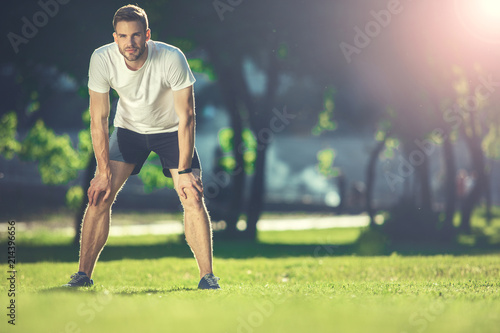Full length portrait of focused sportsman training in park. He is bending forward and putting hands on knees with concentration. Copy space in right side