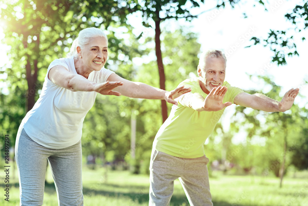 Bending together. Two positive pensioners leading a healthy lifestyle while exercising in a park Bending together. Two positive pensioners leading a healthy lifestyle while exercising in a park