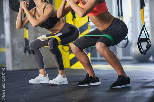 Close up of athletic women in squat together in gym. Couple of fit girls are exercising with resistance band for lower body relief. They are wearing sport clothes and sneakers photo