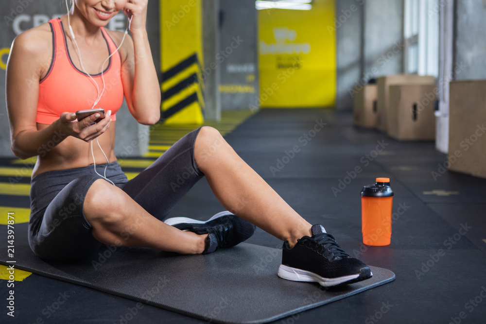 Close up of female legs in sneakers while she is sitting on mat with bottle of water. Slim grinning lady is exercising in fitness room and listening to songs on her smartphone with earphones Close up of female legs in sneakers while she is sitting on mat with bottle of water. Slim grinning lady is exercising in fitness room and listening to songs on her smartphone with earphones