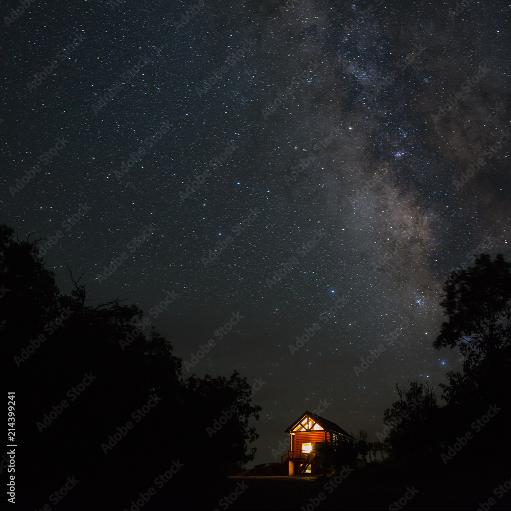 A cabin with it's windows glowing bright under the milky way at night and surrounded by the silhouetted oak trees