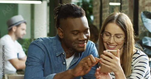 Young multietnical happy couple sitting together at the bar and using smartphone device, taping and scrolling. Close up. photo