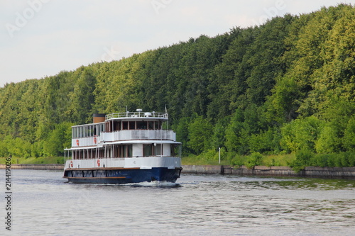 Travel, tourism, water recreation - white tourist ship slowly floating along the concrete coast of the navigable canal in the summer against the green trees on the shore, side view