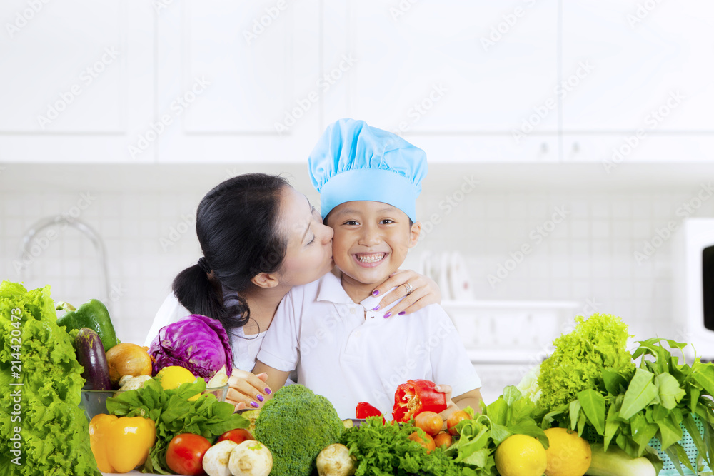 Child kissed by mother with vegetables in kitchen Child kissed by mother with vegetables in kitchen