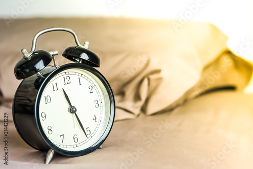 Black cover alarm clock is putting on a bed with pillow behind in brown cover with yellow sun light at late morning