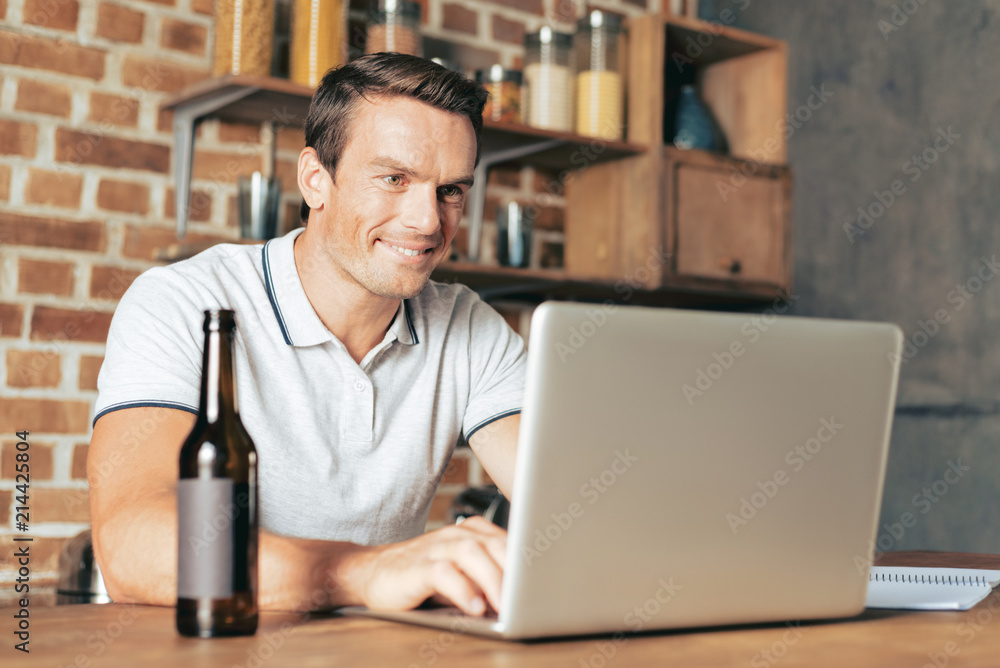 Modern device. Cheerful happy delighted man smiling and using his laptop while having beer Modern device. Cheerful happy delighted man smiling and using his laptop while having beer