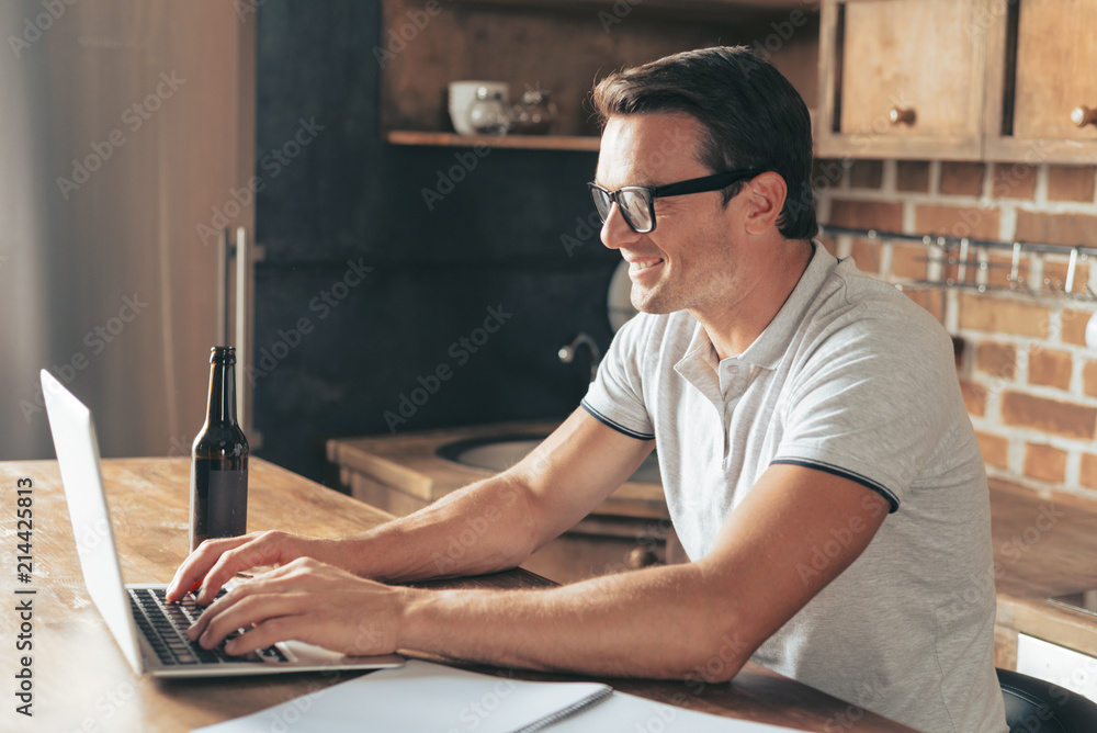 Pleasurable job. Joyful positive handsome man sitting in front of the laptop and typing while working at home