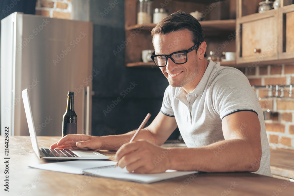 Fototapeta premium For work. Cheerful positive delighted man sitting at the laptop and taking notes while working at home