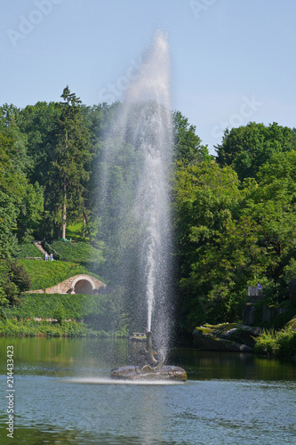 Transparent water fountain striking in the middle of the lake against a background of green nature and slopes in the park with people walking along the paths.