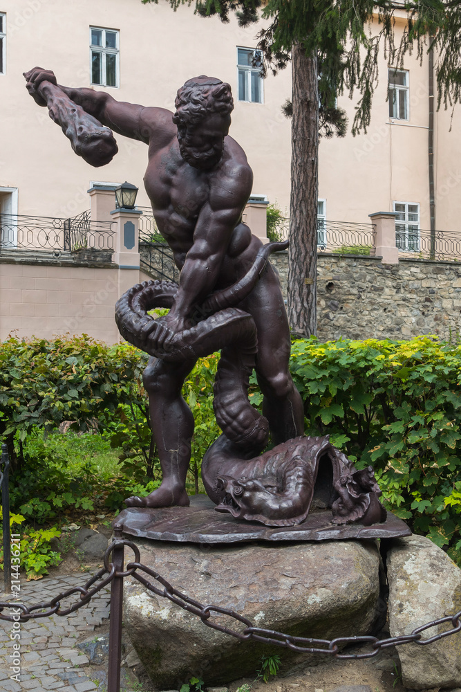 Sculpture depicting a man with a truncheon of a conquering serpent near his feet. Standing on the stones behind the fence with a chain near the green bushes in front of the house.