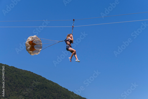 A woman riding along the cable clutching her hands with a stick with rollers tied for back insurance with a small parchute on the background of a green mountain slope and a blue sky.
