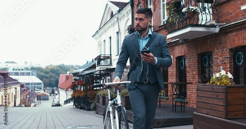 TRACKING Handsome young adult man wearing suit choosing music on his phonee before riding his classic bicycle to work in the morning. 4K UHD 60 FPS SLOW MOTION photo