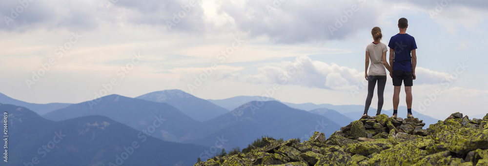 Φωτογραφία Back view of young tourist couple, athletic man and slim girl stand holding hand on rocky mountain top enjoying breathtaking summer mountain panorama