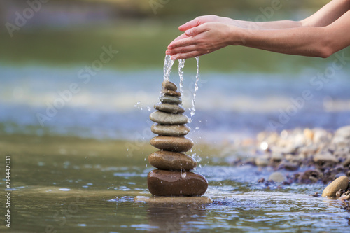 Canvas Print Close-up abstract image of woman hand pouring water on rough natural brown uneven different sizes and form stones balanced like pyramid pile landmark on blurred blue-green copy space background