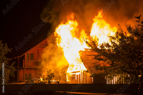 Burning wooden house at night. Bright orange flames and dense smoke from under the tiled roof on dark sky, trees silhouettes and residential neighbor cottage background. Disaster and danger concept.