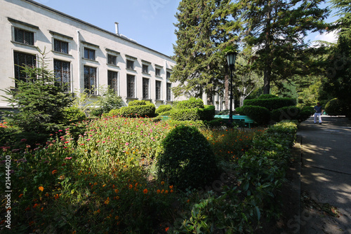 a flowerbed in the shade of trees near the building with barred windows and walking along the path