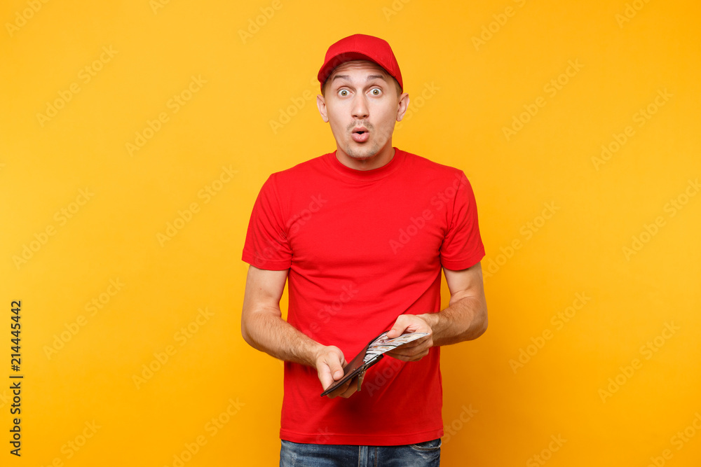 Delivery man in red uniform isolated on yellow orange background. Professional male employee in cap, t-shirt courier or dealer holding bundle lots of dollars, cash money. Service concept. Copy space.