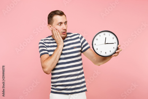 Portrait of shocked sad upset man wearing striped t-shirt holding round clock, copy space isolated on trending pastel pink background. People sincere emotions lifestyle concept. Time is running out.
