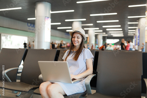 Young smiling traveler tourist woman in hat working on laptop while waiting in lobby hall at international airport. Passenger traveling abroad on weekends getaway. Air travel, flight journey concept.