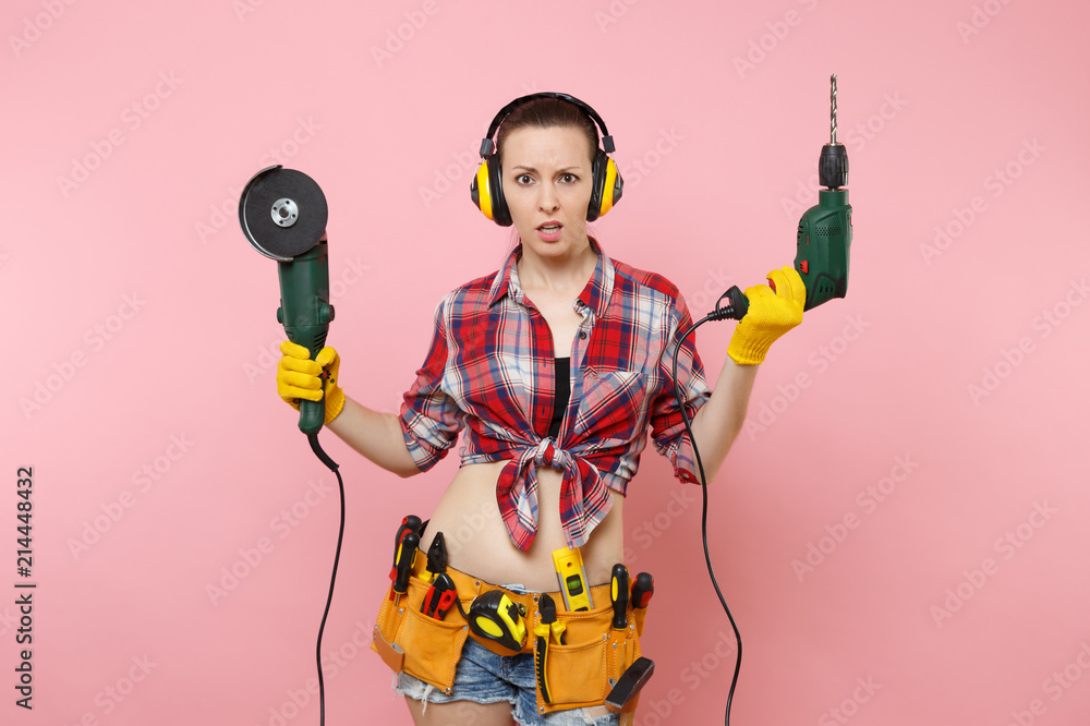 Energy handyman woman in gloves, noise insulated headphones, kit tools belt full of instruments holding power saw electric drill isolated on pink background. Female in male work. Renovation concept. Energy handyman woman in gloves, noise insulated headphones, kit tools belt full of instruments holding power saw electric drill isolated on pink background. Female in male work. Renovation concept.