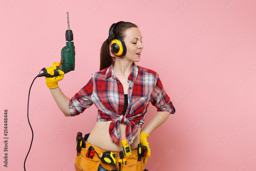 Energy sexy handyman woman in gloves, noise insulated headphones, kit tools  belt full of instruments holding power electric drill isolated on pink  background. Female in male work. Renovation concept. foto de Stock