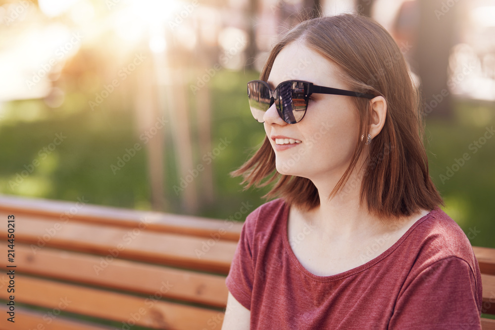 Positive young lovely female with trendy hairstyle, wears fashionable shades, sits on bench in park, enjoys sunny weather during summer time, looks into distance. Outdoor shot of cheerful student Positive young lovely female with trendy hairstyle, wears fashionable shades, sits on bench in park, enjoys sunny weather during summer time, looks into distance. Outdoor shot of cheerful student