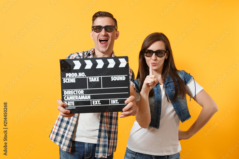 Young fun couple woman man in 3d glasses watching movie film on date holding classic black film making clapperboard pointing index finger up isolated on yellow background. Emotions in cinema concept.