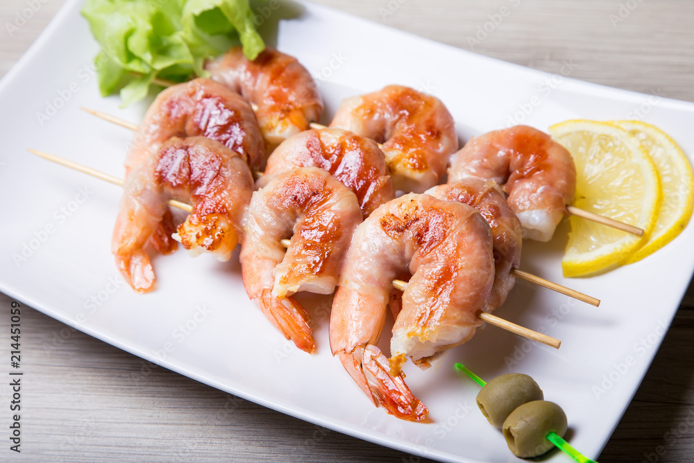 Shrimps grill in prosciutto with lemon and olives on a white plate. Close-up, selective focus. Shrimps grill in prosciutto with lemon and olives on a white plate. Close-up, selective focus.