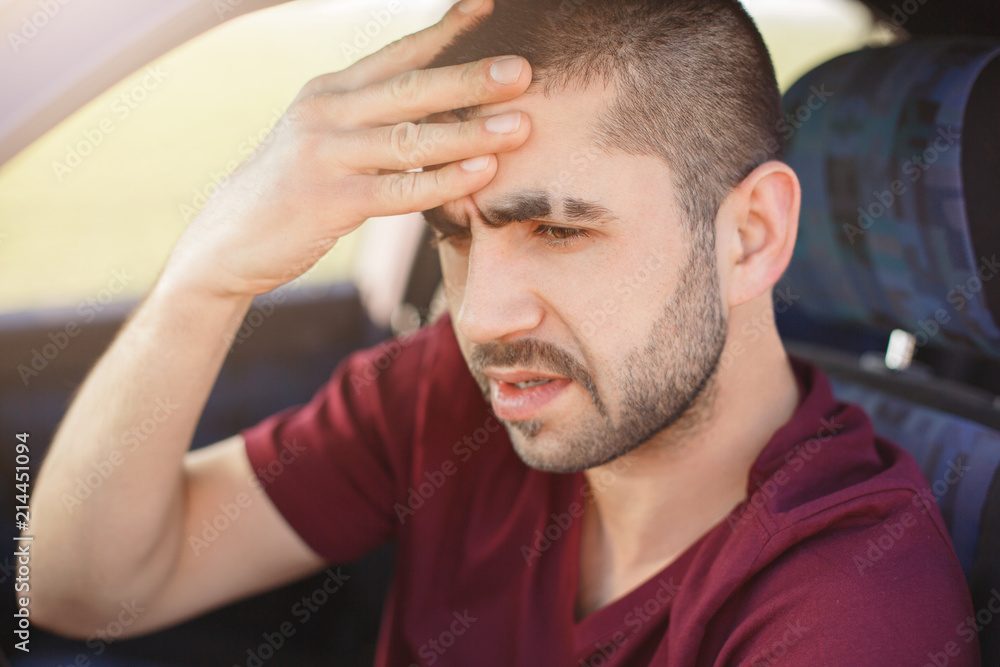 Fatigue male driver keeps hand on forehead, has headache after being long time in car, dressed casually, thinks about future trip. Professional man drives automobile. People and transport concept Fatigue male driver keeps hand on forehead, has headache after being long time in car, dressed casually, thinks about future trip. Professional man drives automobile. People and transport concept