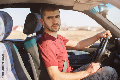 Serious handsome bearded male driver poses in his car, rides auto, uses safety belt, being experienced, looks confidently at camera. Man instructor teaches to drive, dressed in casual red t shirt