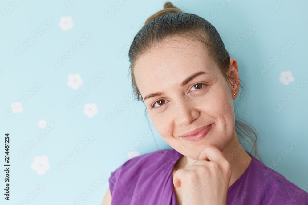 Fototapeta premium Close up portrait of pretty delighted young woman holds chin, looks positively at camera, enjoys having weekend, dressed in purple clothes, stands against blue wall. People and lifestyle concept