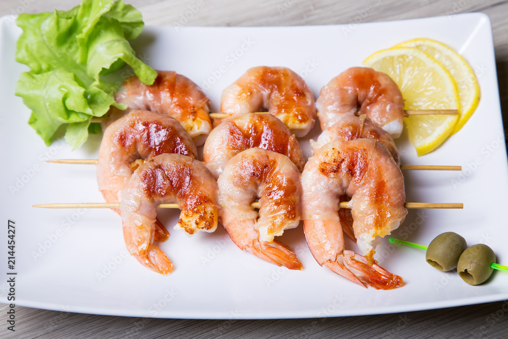 Shrimps grill in prosciutto with lemon and olives on a white plate. Close-up, selective focus.