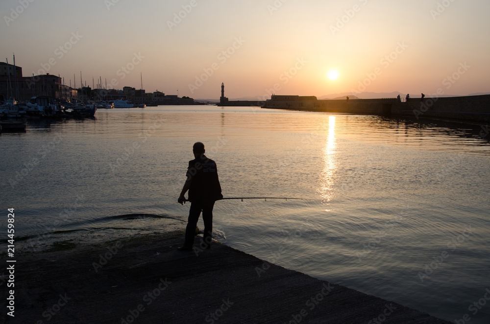 Fototapeta premium Silhouette of fisherman with lighthouse and sunset on background. Sea sunset, sunset on the sea in Greece, Chania. Golden orange sunset