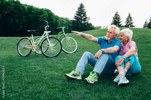 happy senior couple sits on the grass, enjoying the rest after riding bicycles. Bicycles stand next to them. Active rest retired
